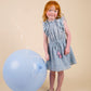 A young girl in the Early Sunday Girls’ Florence Dress, made from tonal floral blue Swiss dobby, stands beside a large blue balloon against a simple beige background.