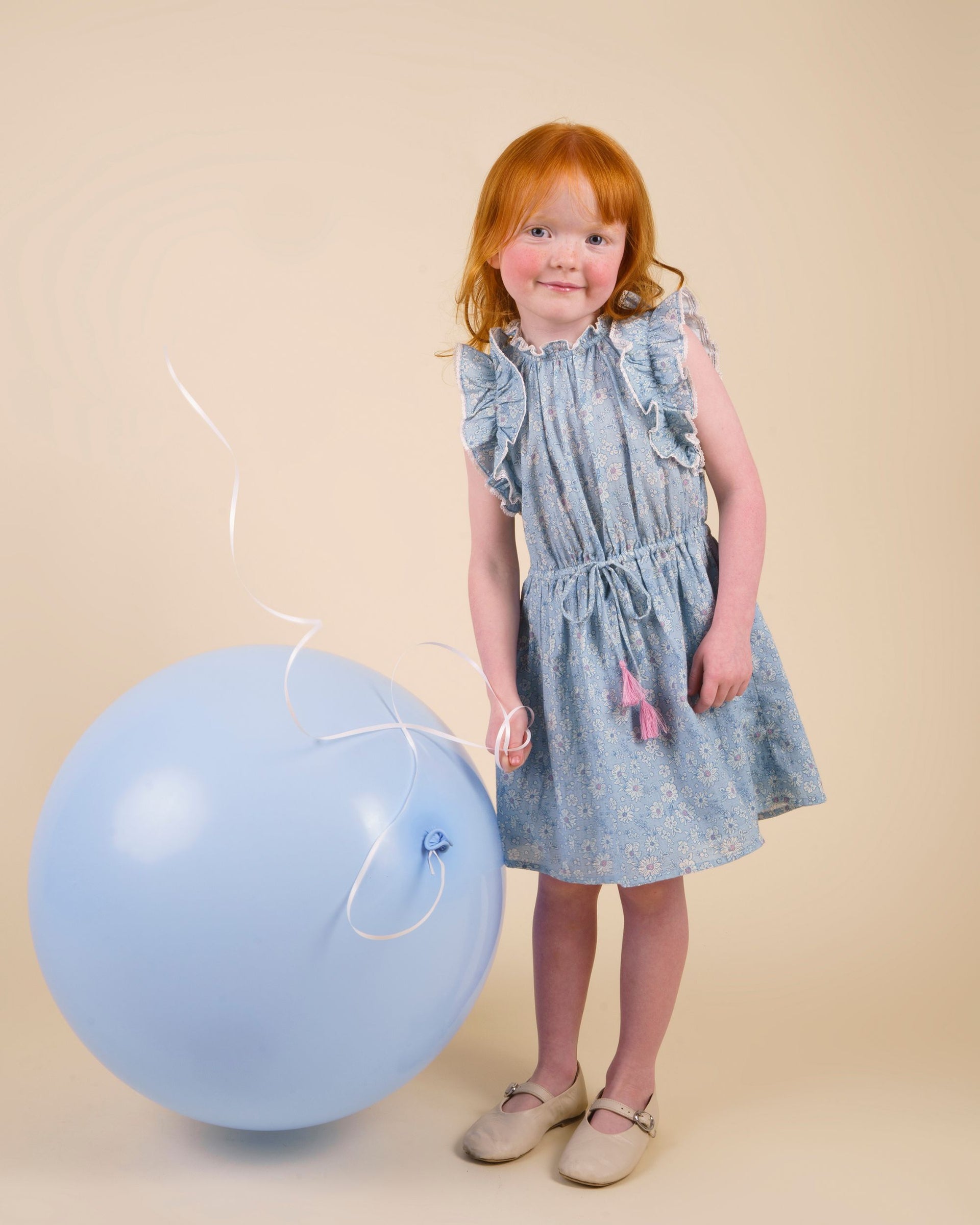 A young girl in the Early Sunday Girls’ Florence Dress, made from tonal floral blue Swiss dobby, stands beside a large blue balloon against a simple beige background.