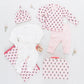 A flat lay of Pink Chicken's Nesting Box Baby Take Home Set in organic pima cotton features a hat, two long-sleeve tops, pants, white onesie, floral blanket, small bag, pacifier, and decorative flowers arranged around them.