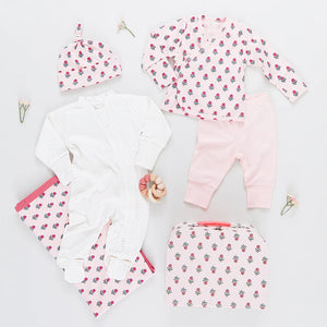 A flat lay of Pink Chicken's Nesting Box Baby Take Home Set in organic pima cotton features a hat, two long-sleeve tops, pants, white onesie, floral blanket, small bag, pacifier, and decorative flowers arranged around them.