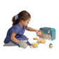 A child in a blue dress, possibly enjoying a delightful toddler birthday gift, plays with the Manhattan Toys Forest Tales Baking Set by Manhattan Toy, holding a piece of pretend bread. A blue toy oven and various wooden play kitchen accessories are arranged in front of the child.