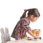 A young child, wearing a floral dress with a ribbon in her hair, plays with a Tenderleaf Fruity Blender from Tender Leaf Toys at her small play kitchen set. There is a wooden chair with a bunny face next to her, completing the scene of her imaginative smoothie stall.