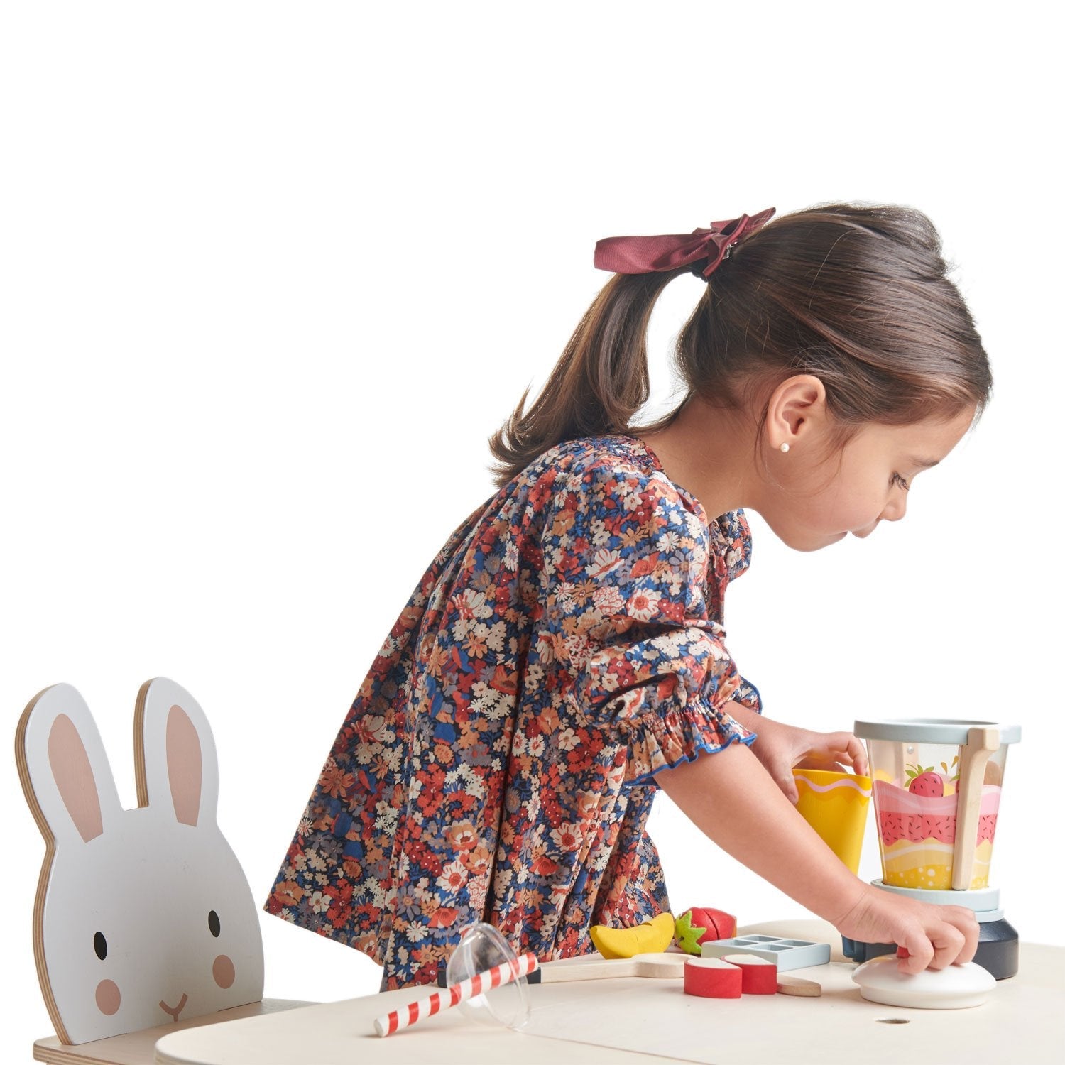 A young child, wearing a floral dress with a ribbon in her hair, plays with a Tenderleaf Fruity Blender from Tender Leaf Toys at her small play kitchen set. There is a wooden chair with a bunny face next to her, completing the scene of her imaginative smoothie stall.