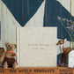 A beige book titled "Write To Me Funny Things You Say Oatmeal" is placed upright on a brick-patterned surface, resembling a charming journal of kids' quotes. A stuffed bear and reindeer are positioned on either side, adding a whimsical touch.