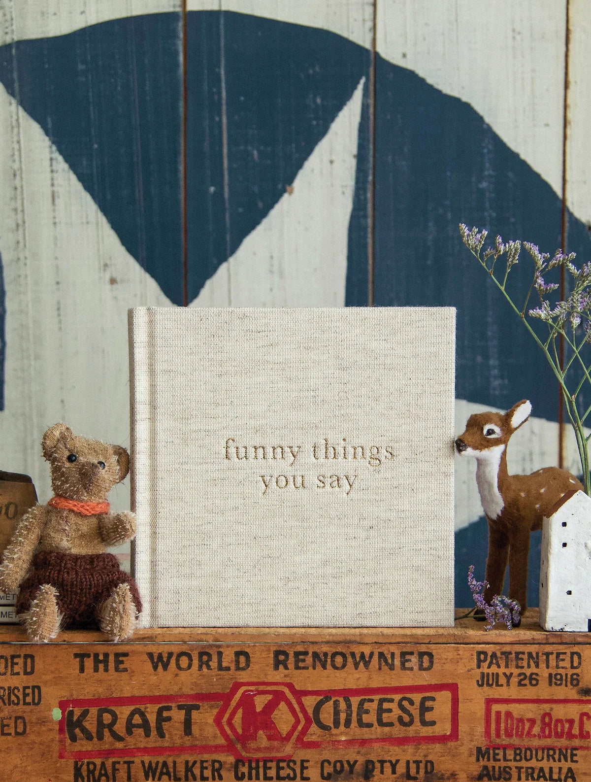 A beige book titled "Write To Me Funny Things You Say Oatmeal" is placed upright on a brick-patterned surface, resembling a charming journal of kids' quotes. A stuffed bear and reindeer are positioned on either side, adding a whimsical touch.