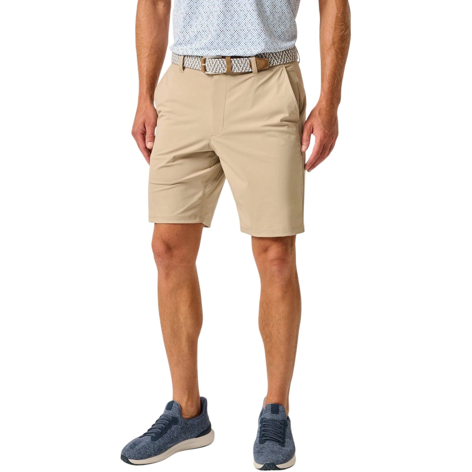 A man wearing a patterned belt, light blue shirt, Johnnie-O Fusionn Short in beige, and blue sneakers stands against a white background.