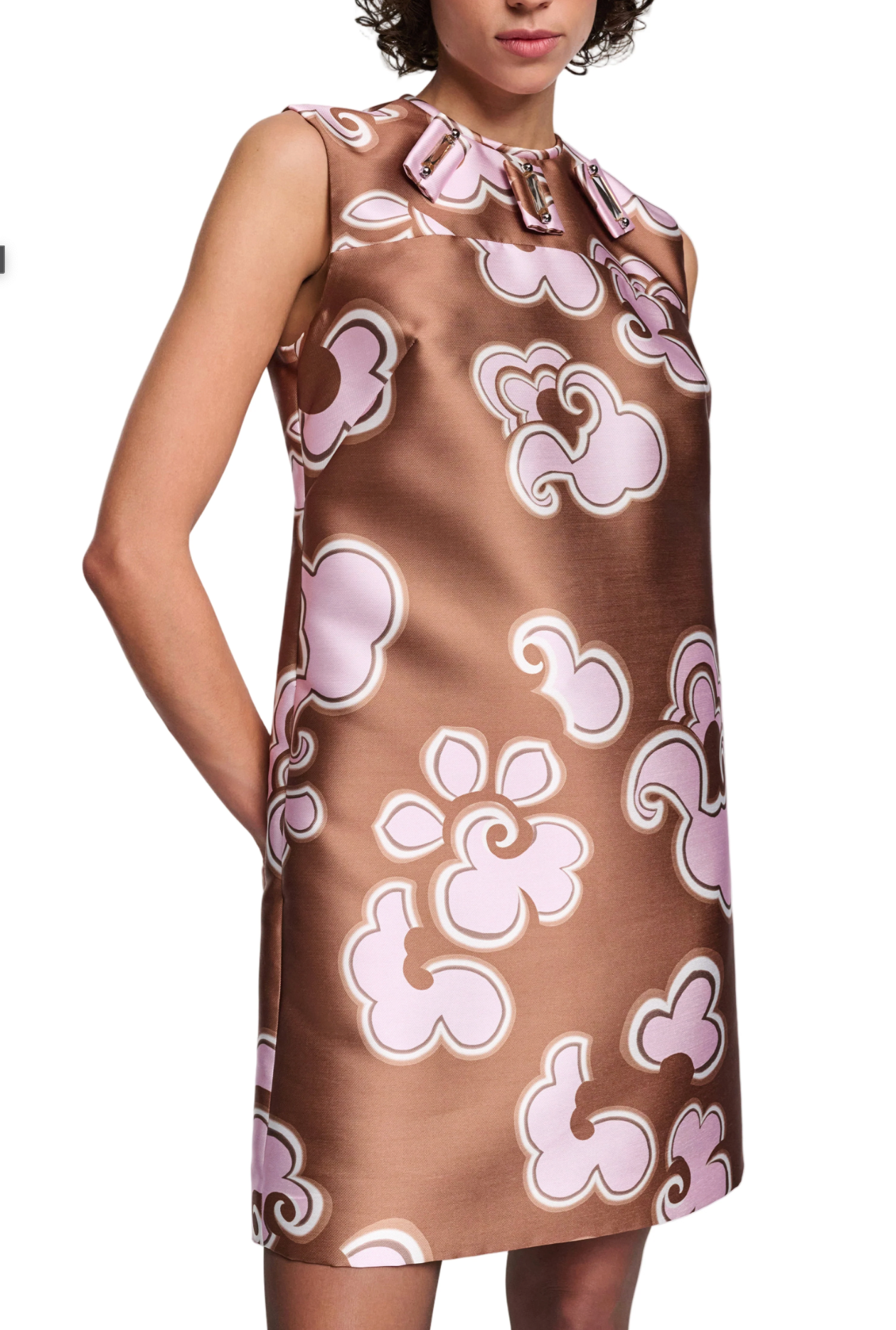 A woman wears the Dorothee Schumacher Floral Statement Dress, a sleeveless brown satin shift with a pink floral print, bow neckline, handmade embellishments, and pockets where her hands rest casually.