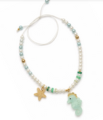 Introducing the Seahorse Green Necklace by Lillies & Roses, a handcrafted piece featuring white, blue, and gold beads, beautifully accented with a gold starfish charm and a light blue seahorse pendant on a white adjustable cord.