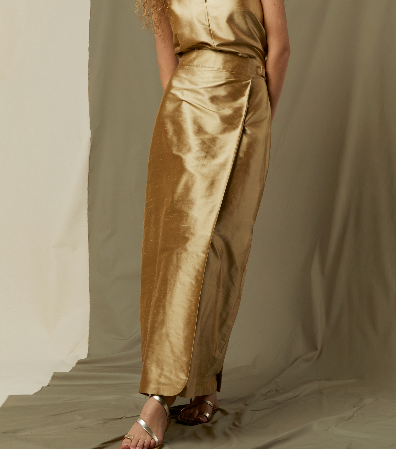 Against a beige backdrop, a person wears the Ann Mashburn Renata Silk Shantung Wrap Skirt with a matching sleeveless top and sandals. Their head and feet are partially cropped from the frame.