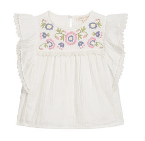 The Girls’ Maiya Blouse by Louise Misha is a white short-sleeve cotton gauze blouse featuring ruffled lace trim and multicolored floral embroidery on the chest.