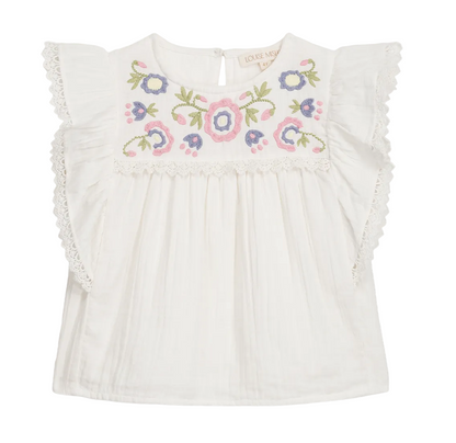 The Girls’ Maiya Blouse by Louise Misha is a white short-sleeve cotton gauze blouse featuring ruffled lace trim and multicolored floral embroidery on the chest.