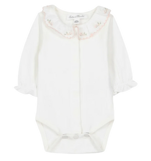 The Tartine et Chocolat Baby Girls’ Ruffled Collar Bodysuit is a white long-sleeve onesie crafted from soft ecru cotton, featuring ruffled cuffs, convenient snap closures, and an embroidered scalloped collar adorned with delicate floral details. This charming bodysuit blends comfort and elegance for a sweet, timeless look.