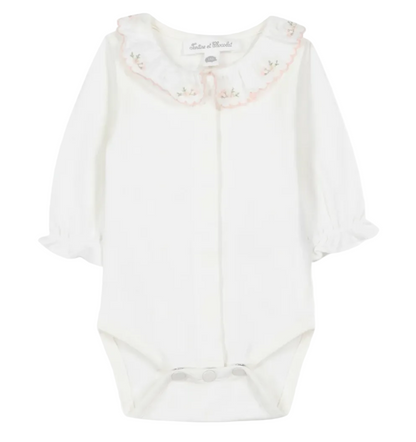 The Tartine et Chocolat Baby Girls’ Ruffled Collar Bodysuit is a white long-sleeve onesie crafted from soft ecru cotton, featuring ruffled cuffs, convenient snap closures, and an embroidered scalloped collar adorned with delicate floral details. This charming bodysuit blends comfort and elegance for a sweet, timeless look.