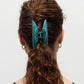 A person with curly brown hair styled in a low ponytail, secured with the Machete Grande Heirloom Claw in teal Italian acetate, is shown from the back. The person is wearing gold hoop earrings and a black top.