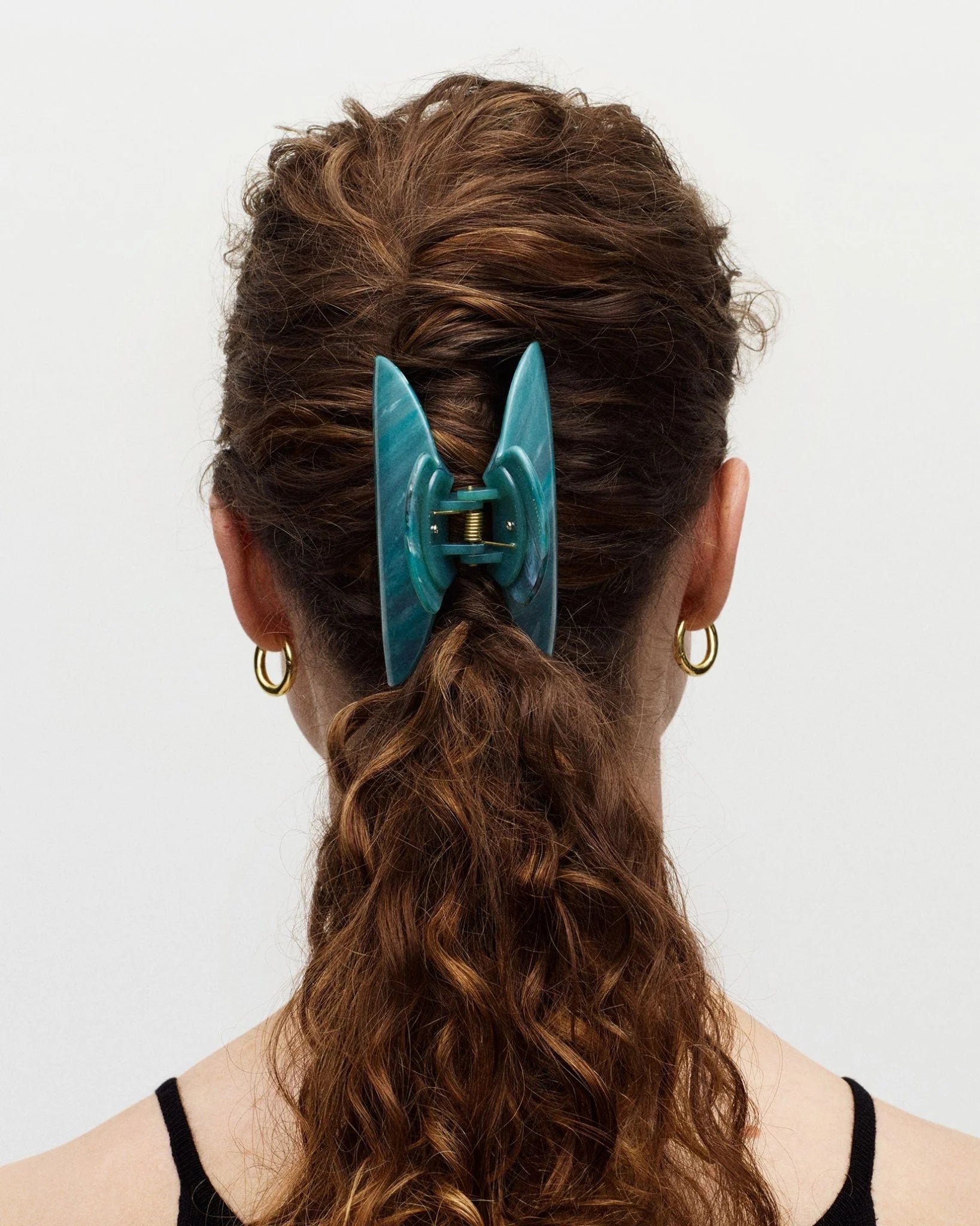 A person with curly brown hair styled in a low ponytail, secured with the Machete Grande Heirloom Claw in teal Italian acetate, is shown from the back. The person is wearing gold hoop earrings and a black top.