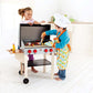 Two children are playing with a Hape Gourmet Grill. The girl is handing a patty to the boy, who is wearing a chef's hat and apron. The scene takes place in a bright, modern kitchen with white cabinets, and the Hape Gourmet Grill features double-sided grates for versatile cooking fun.