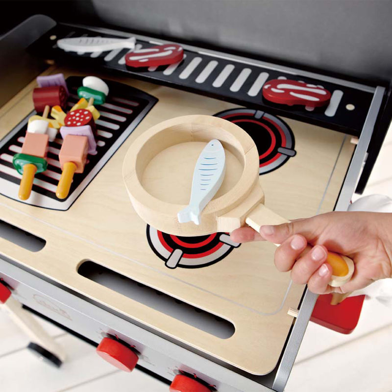 A child's hand holding a toy frying pan with a wooden fish, next to a Hape Gourmet Grill with double-sided grates and skewered wooden food items from the ShishKabob Basics Set.