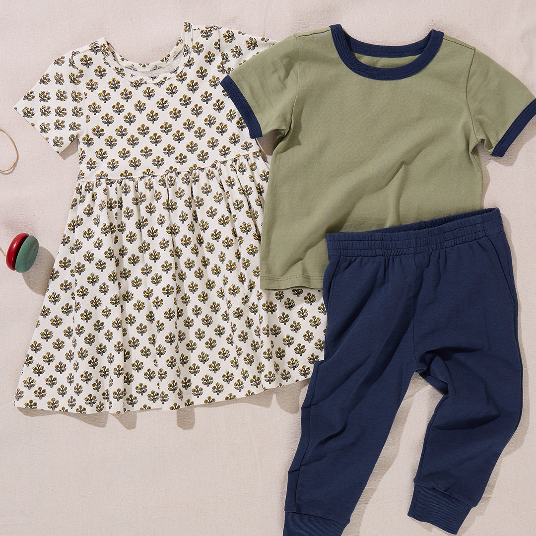 A patterned dress, a green short-sleeve shirt with navy trim, and a pair of Coop Kids PC Boys’ Jax Jogger pants in soft French Terry are laid out on a beige surface next to a small toy.