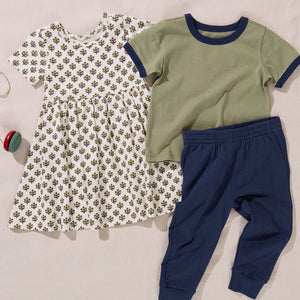 A patterned dress, a green short-sleeve shirt with navy trim, and a pair of Coop Kids PC Boys’ Jax Jogger pants in soft French Terry are laid out on a beige surface next to a small toy.
