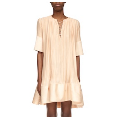 A person wearing the Lanvin Short Sleeve Cascade Dress—a loose, knee-length light beige charmeuse dress with short sleeves, a V-neckline, and a ruffled hem.