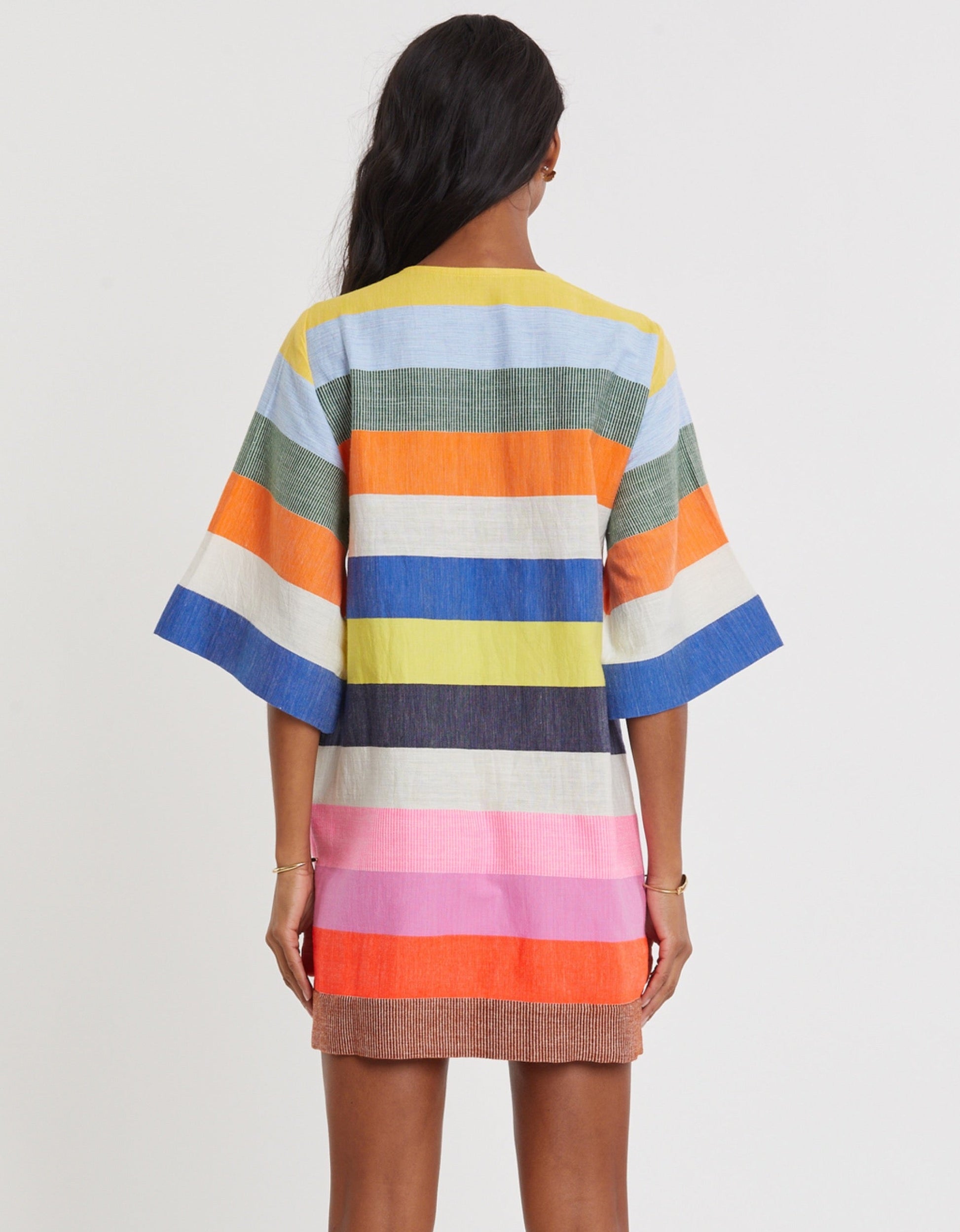 A person stands facing away, wearing the Lemlem Belkis Caftan—a knee-length, hand-woven garment with wide horizontal stripes in blue, orange, green, pink, and yellow.