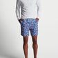 A person stands against a plain background wearing a long-sleeve, white quarter-zip shirt, Peter Millar Hammerhead Camo Swim Trunk made of quick-drying, four-way stretch fabric, and flip-flops. Their hands are in their shorts pockets.