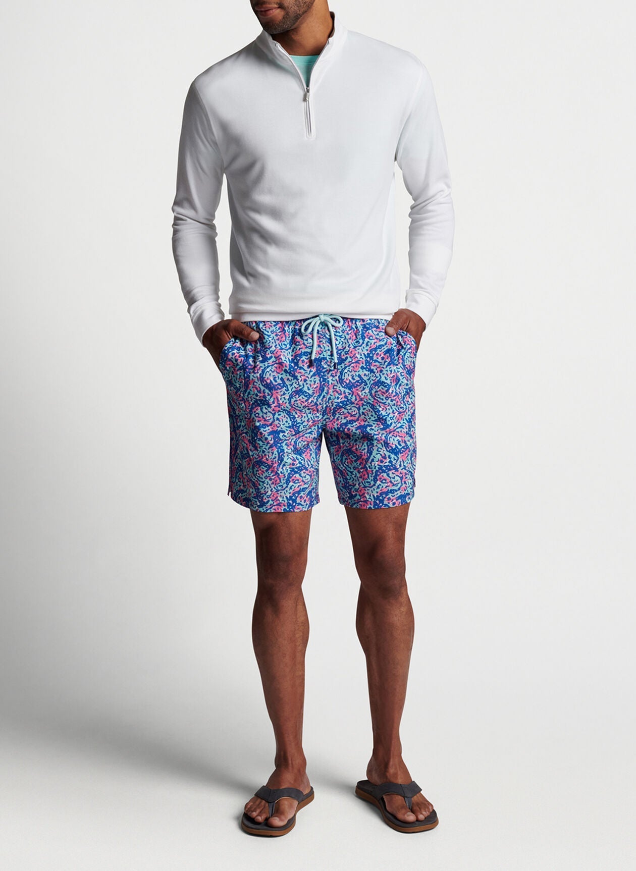 A person stands against a plain background wearing a long-sleeve, white quarter-zip shirt, Peter Millar Hammerhead Camo Swim Trunk made of quick-drying, four-way stretch fabric, and flip-flops. Their hands are in their shorts pockets.