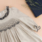A light gray Kidiwi Baby Girls' and Girls' Hars Organic Cotton Twill Smocked Dress with black embroidery is displayed next to a dark blue book and green sprigs on a beige surface.