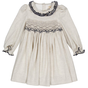 The Kidiwi Baby Girls' and Girls' Hars Organic Cotton Twill Smocked Dress features a cream color, long sleeves, ruffled cuffs, a gathered skirt, and black embroidery on the collar and chest.