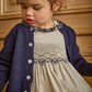 A young child with curly hair wears the Kidiwi Baby Girls' and Girls' Hars Organic Cotton Twill Smocked Dress, featuring embroidered details, paired with a navy cardigan, standing before a wooden backdrop.