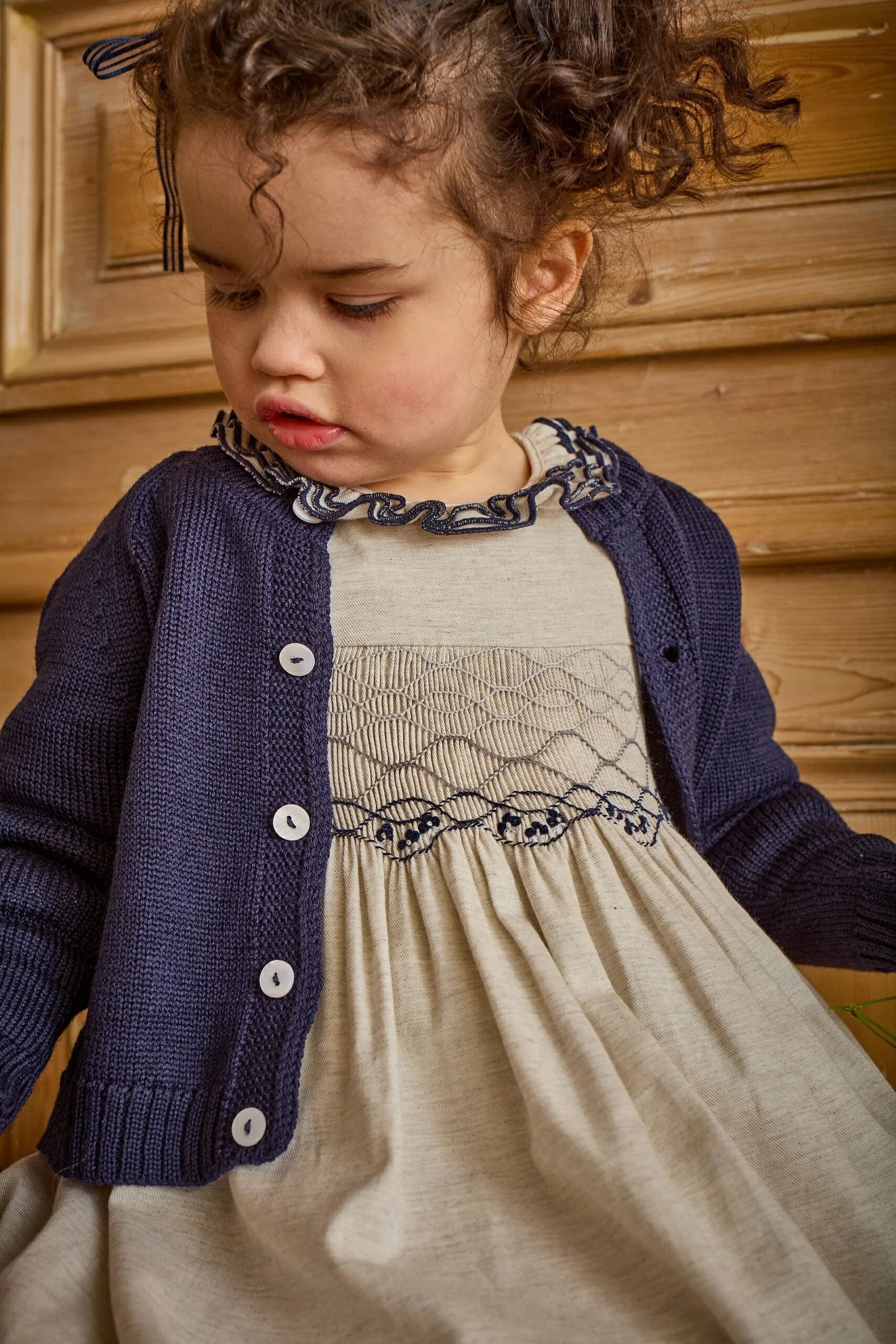A young child with curly hair wears the Kidiwi Baby Girls' and Girls' Hars Organic Cotton Twill Smocked Dress, featuring embroidered details, paired with a navy cardigan, standing before a wooden backdrop.