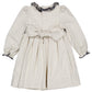 The Kidiwi Baby Girls' and Girls' Hars Organic Cotton Twill Smocked Dress features long sleeves, a beige hue, black ruffle trim on the collar and cuffs, a buttoned back, and a large waist bow.