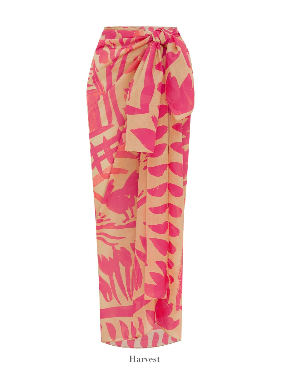 The Soleil Soleil Square Pareo by Soleil Soleil is a long wrap-around skirt featuring a vibrant pink and beige leaf pattern. The waistband is tied in a bow, making it perfect as an organic cotton beach cover-up. Text beneath reads "Harvest.