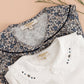 A folded Kidiwi Baby Girls' Hélena Blouse with Navy Embroidery is displayed on a peach background alongside dried flowers.