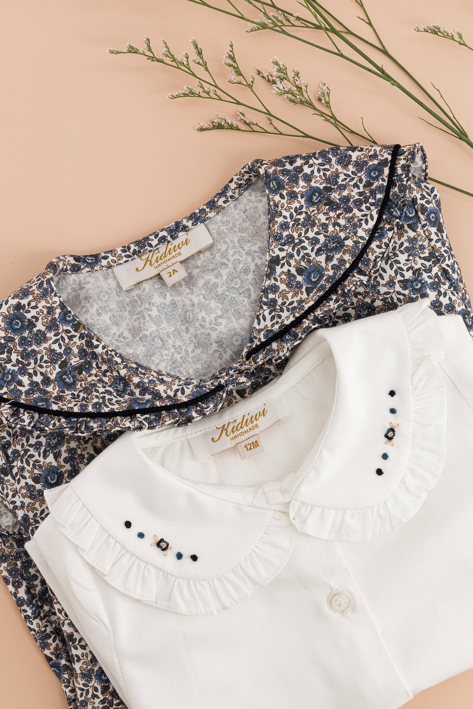 A folded Kidiwi Baby Girls' Hélena Blouse with Navy Embroidery is displayed on a peach background alongside dried flowers.