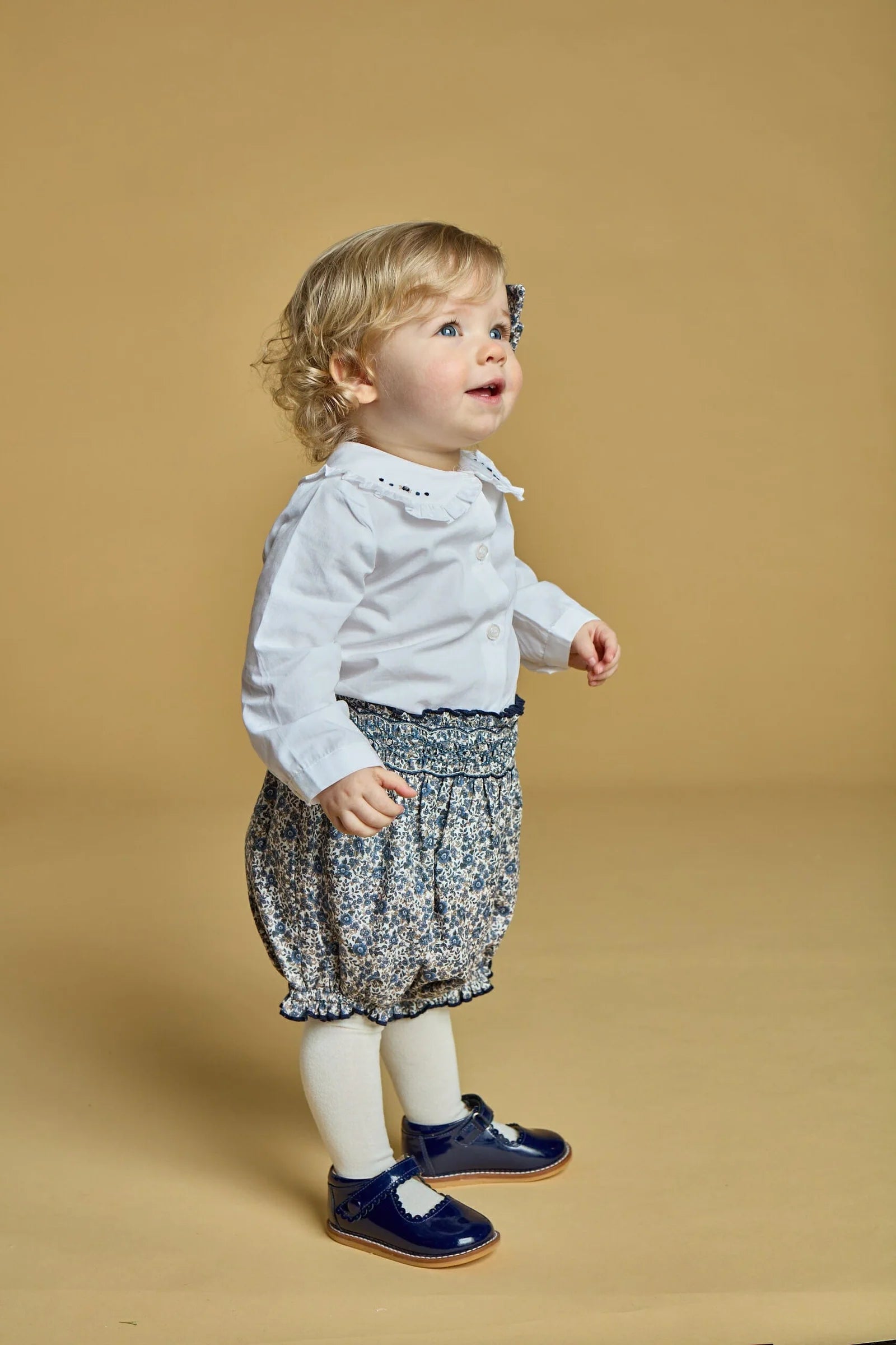 A toddler with blonde curly hair stands on a beige background, wearing the Kidiwi Baby Girls' Hélena Blouse with Navy Embroidery, floral shorts, white tights, and blue shoes.