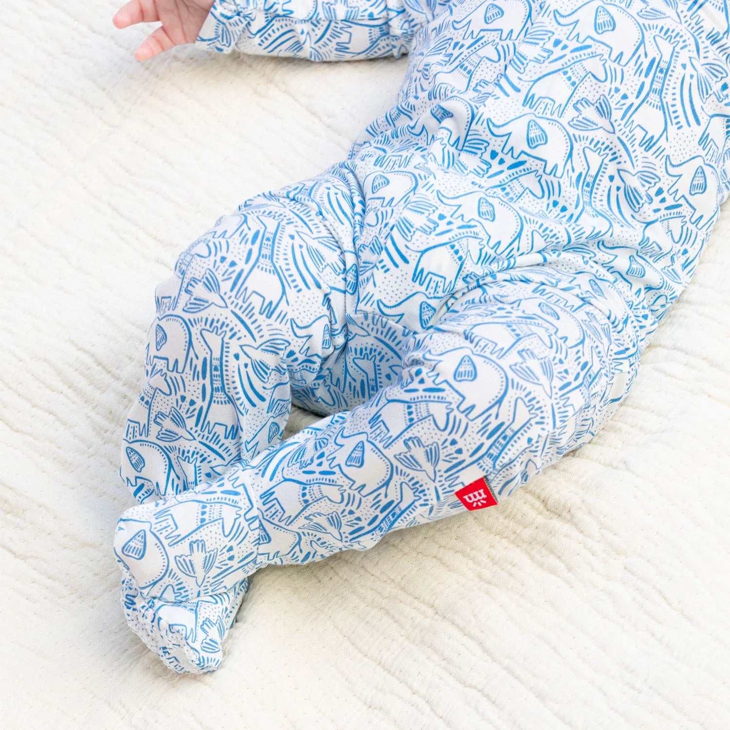 A baby wearing Magnetic Me's Baby' Heards of Feather Modal Footie with feet lies on a cream-colored textured blanket.