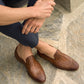 Seated on a stone surface, a person wears stylish dark pants and Magnanni's Herrera Woven Loafers in brown, their hands resting gently on their knees.