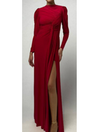 A person wears the Monique Lhuillier Jewel Neck Long Sleeve Draped Gown in red matte jersey, featuring a draped bodice, gathered detailing, and a high thigh slit, standing against a plain background.