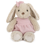 Bettie Bunny - Pink by Mon Ami is a beige plush bunny with floppy ears, dressed in a pink dress and matching bow, sitting facing forward against a white background. This soft stuffed animal is perfect for cuddles and display.