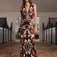 A woman stands in a stable aisle wearing the Johanna Ortiz Horizonte Mistico Midi Dress, a sleeveless, midi-length design featuring the Equine Mola Tiles motif with a deep V-neck, paired with sandals, a crossbody bag, and a necklace.