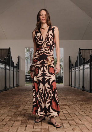 A woman stands in a stable aisle wearing the Johanna Ortiz Horizonte Mistico Midi Dress, a sleeveless, midi-length design featuring the Equine Mola Tiles motif with a deep V-neck, paired with sandals, a crossbody bag, and a necklace.