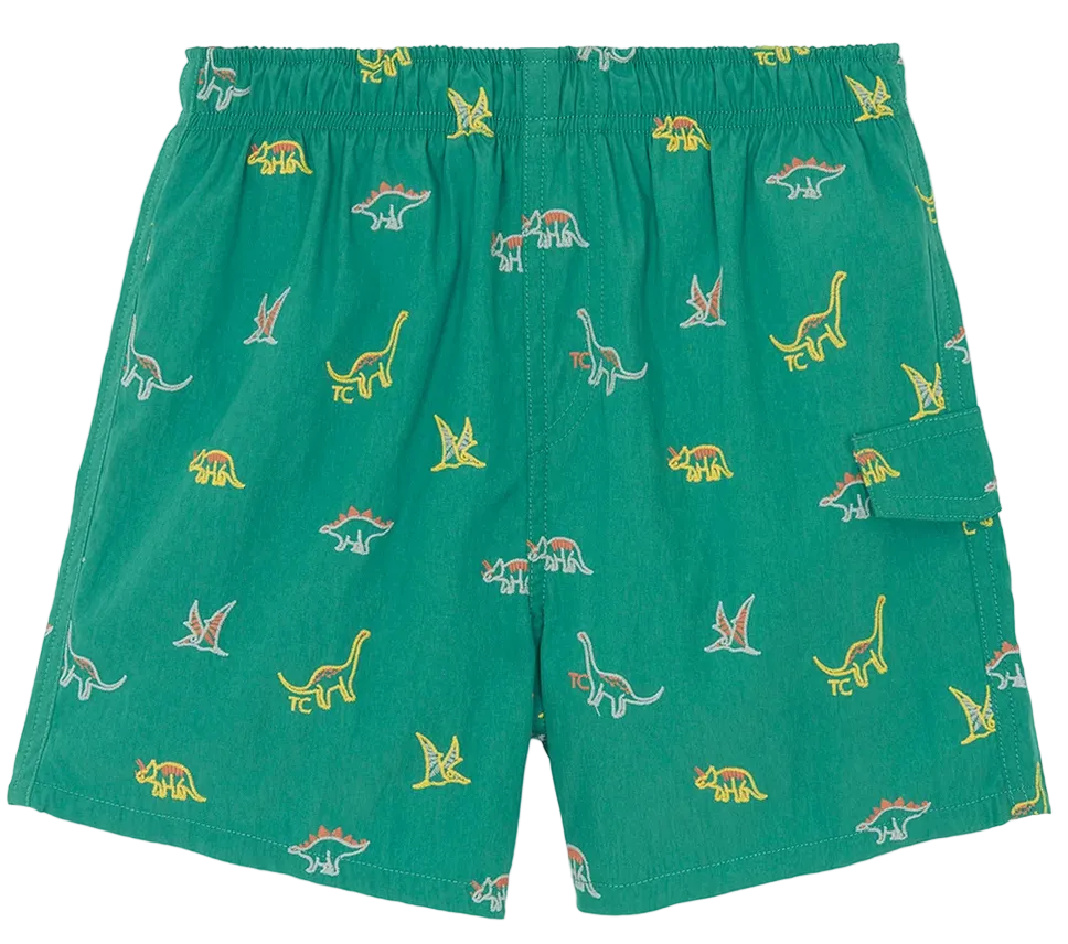 The Boys’ Printed Swim Short by Tartine et Chocolat features a vibrant dinosaur print on green fabric, an elastic waistband for added comfort, and a handy side pocket, shown laid flat against a white background.