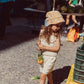 A young girl in a beige outfit and a vibrant Louise Misha Lajik Hat holds vegetables at an outdoor market, surrounded by crates and blurred people in the background.