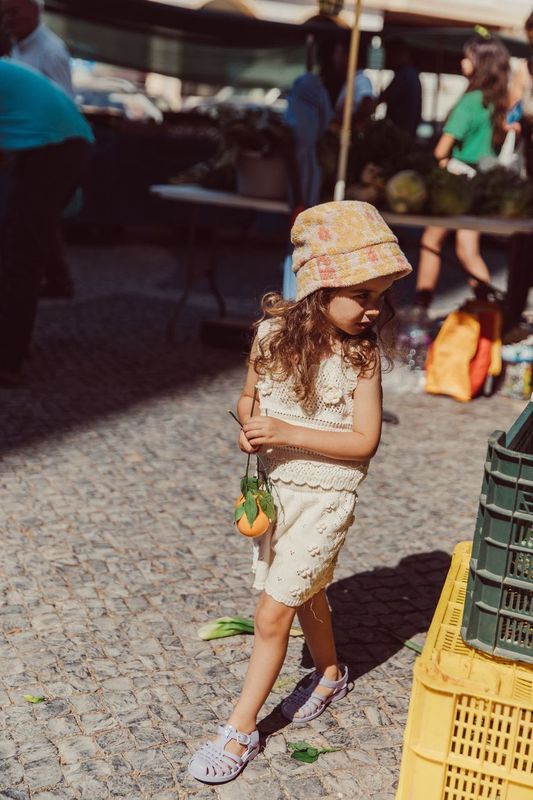 A young girl in a beige outfit and a vibrant Louise Misha Lajik Hat holds vegetables at an outdoor market, surrounded by crates and blurred people in the background.