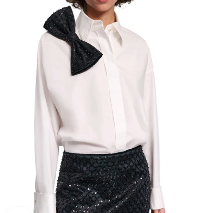 A person wears the Dorothee Schumacher Poplin Power Bow Blouse, featuring an oversized cotton poplin design with a large black bow on the shoulder, paired with a black sequined skirt.