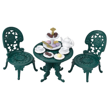 The Calico Critters Tea and Treats Set features two ornate green chairs and a matching garden table, charmingly arranged with tea party accessories like a white teapot, teacups, plates, and a tray of assorted pastries and sweets.