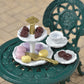 Display your favorite tea party accessories with the Calico Critters Tea and Treats Set, featuring a two-tiered tray filled with assorted sweets, a gold serving knife, and a green ornate garden table complete with teacup, chocolates, and a matching chair in the background—the perfect addition to any whimsical gathering.