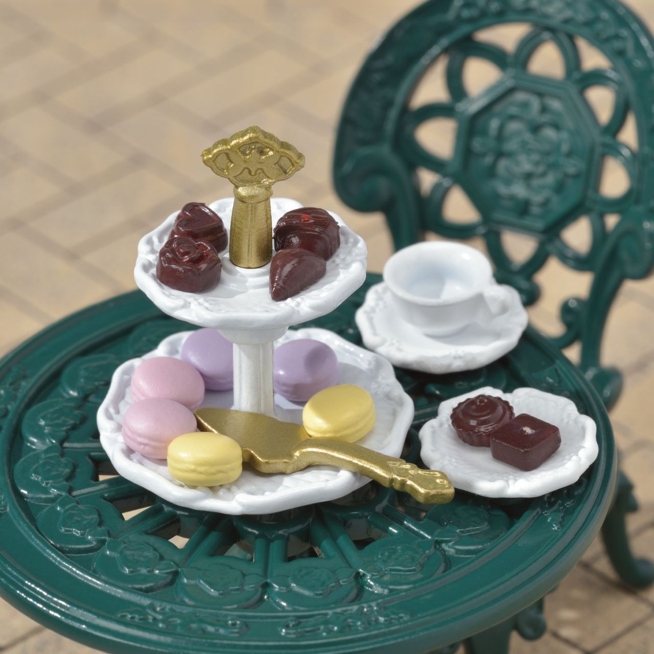 Display your favorite tea party accessories with the Calico Critters Tea and Treats Set, featuring a two-tiered tray filled with assorted sweets, a gold serving knife, and a green ornate garden table complete with teacup, chocolates, and a matching chair in the background—the perfect addition to any whimsical gathering.