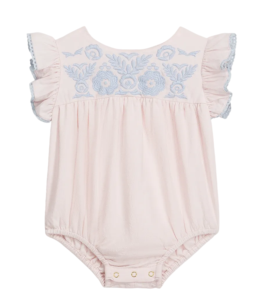 The Louise Misha Baby Girls’ Nusha Romper is crafted from soft cotton gauze in a light pink hue, featuring short ruffled sleeves, an embroidered yoke with delicate blue floral detailing on the chest, and convenient snap closures at the bottom.